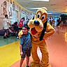 child, boy, mascot, pluto, costume, indoor, cartoon_art, wall_art, people, shoes, shorts, tshirt, smile, family, stroller, backpack, floor, ceiling, lighting, happy