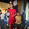 child, boy, football, soccer, cristiano_ronaldo, wax_figure, sports_uniform, red_jersey, football_ball, museum, exhibit, indoor, portrait, smiling, surprised_expression, visitors, sports_fan, photograph, colorful_background, standing