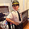 child, boy, pilot_costume, uniform, pilot_hat, smiling, portrait, indoors, bedroom, crib, bed, furniture, tie, white_shirt, black_pants, wood_floor, home, posing, costume, face