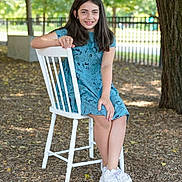Eva participe au concours pour gagner de l'argent avec cette photo : child, girl, smiling, blue_dress, white_chair, outdoor, tree, leaves, mulch, sneakers, casual, portrait, nature, fence, daylight, happy, seated, young, person, park