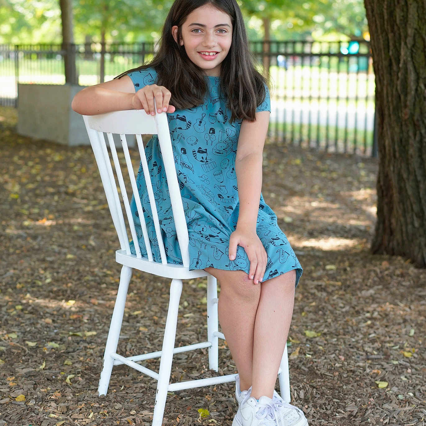Eva participe au concours pour gagner de l'argent avec cette photo : child, girl, smiling, blue_dress, white_chair, outdoor, tree, leaves, mulch, sneakers, casual, portrait, nature, fence, daylight, happy, seated, young, person, park