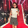 Eliana is registered to the contest to win money with this photo: child, girl, diploma, certificate, glasses, white_shirt, black_skirt, floral_pattern, red_carpet, backdrop, paparazzi, lights, indoors, smile, standing, shoes, socks, long_hair, celebration, achievement