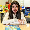 Eva joined the competition — help win amazing prizes! books, braces, brown_hair, child, classroom, desk, educational_materials, girl, indoors, learning, long_hair, orange_ball, photograph, portrait, school, sitting, smiling, student, sweater, table