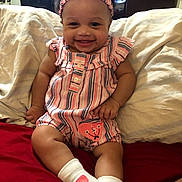 Mia is registered to the contest to win money with this photo: baby, child, smiling, headband, striped_clothing, socks, heart_pattern, bed, pillow, blanket, indoor, happy, person, cute, infant, portrait, seated, home, skin, face