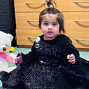 Rijak Kaur joined the competition — help win amazing prizes! toddler, child, black_dress, sparkles, barefoot, carpet, toy, teddy_bear, pink_ribbon, dresser, wooden_furniture, hair_tied_up, indoor, curious_expression, sitting, cute, young_child, face, person, playful