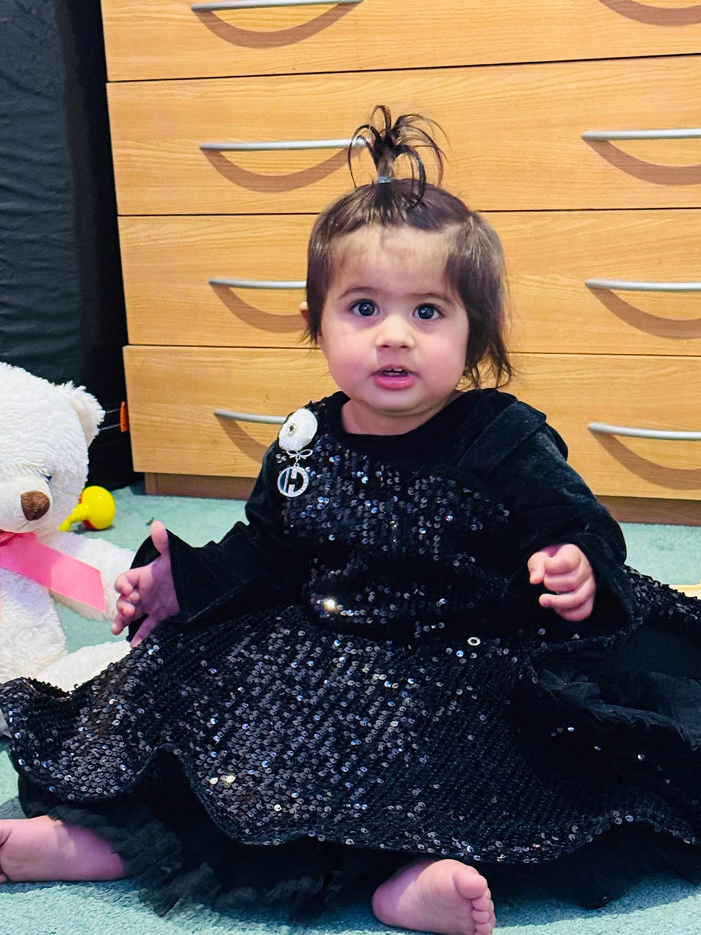 Rijak Kaur joined the competition — help win amazing prizes! toddler, child, black_dress, sparkles, barefoot, carpet, toy, teddy_bear, pink_ribbon, dresser, wooden_furniture, hair_tied_up, indoor, curious_expression, sitting, cute, young_child, face, person, playful