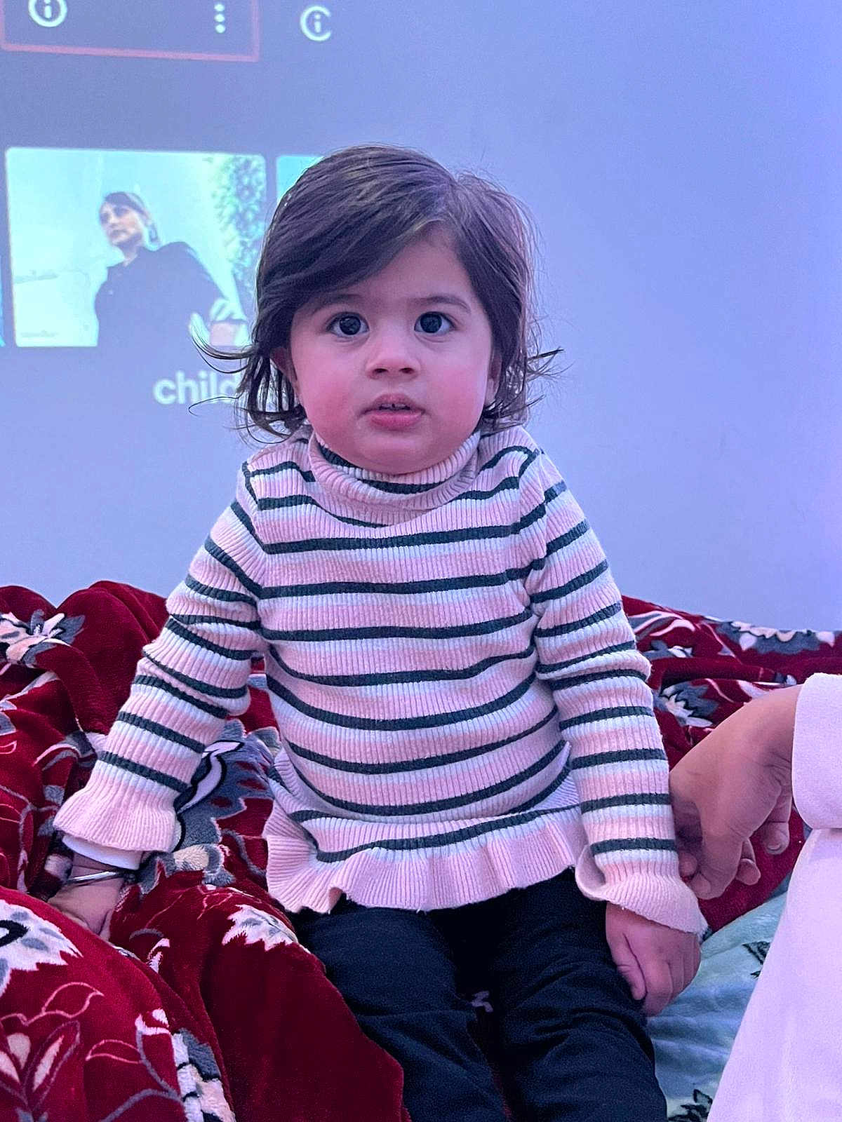 Rijak Kaur joined the competition — help win amazing prizes! child, toddler, striped_sweater, indoor, blanket, patterned_blanket, hand_holding, curly_hair, big_eyes, sitting, person, portrait, casual_clothing, young_child, cozy, soft_lighting, curious_expression, face, home, comfort