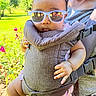 baby, sunglasses, baby_carrier, adult, outdoor, greenery, flowers, tongue_out, casual_clothing, sunlight, person, child, nature, summer, reflection, closeup, cute, playful, portrait, happy