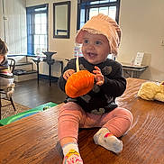 Avis is registered to the contest to win money with this photo: baby, child, smiling, sitting, hat, bonnet, toy, pumpkin, indoor, table, wood, pink_pants, socks, face, person, window, furniture, room, happy, cozy
