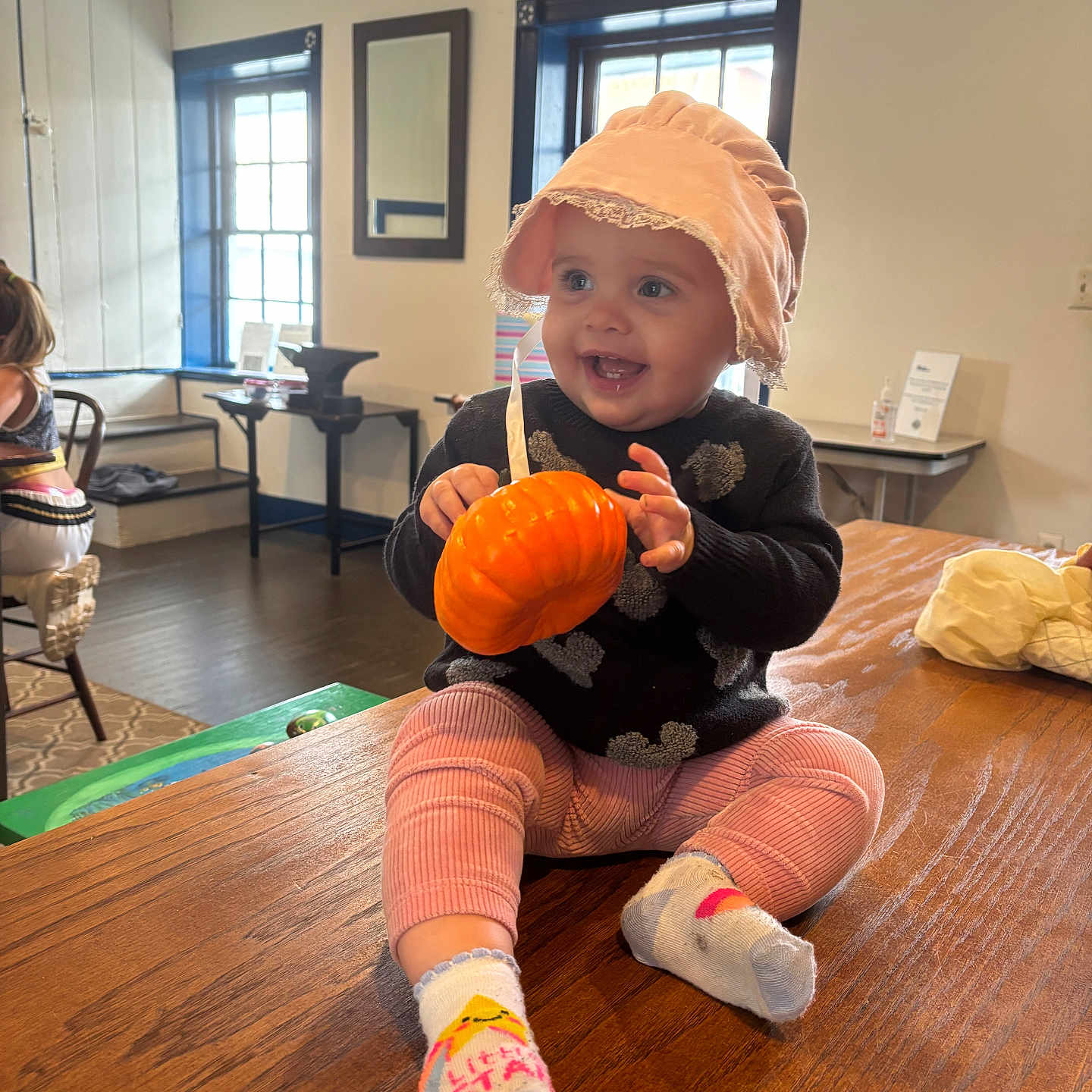 Avis is registered to the contest to win money with this photo: baby, bonnet, child, cozy, face, furniture, happy, hat, indoor, person, pink_pants, pumpkin, room, sitting, smiling, socks, table, toy, window, wood