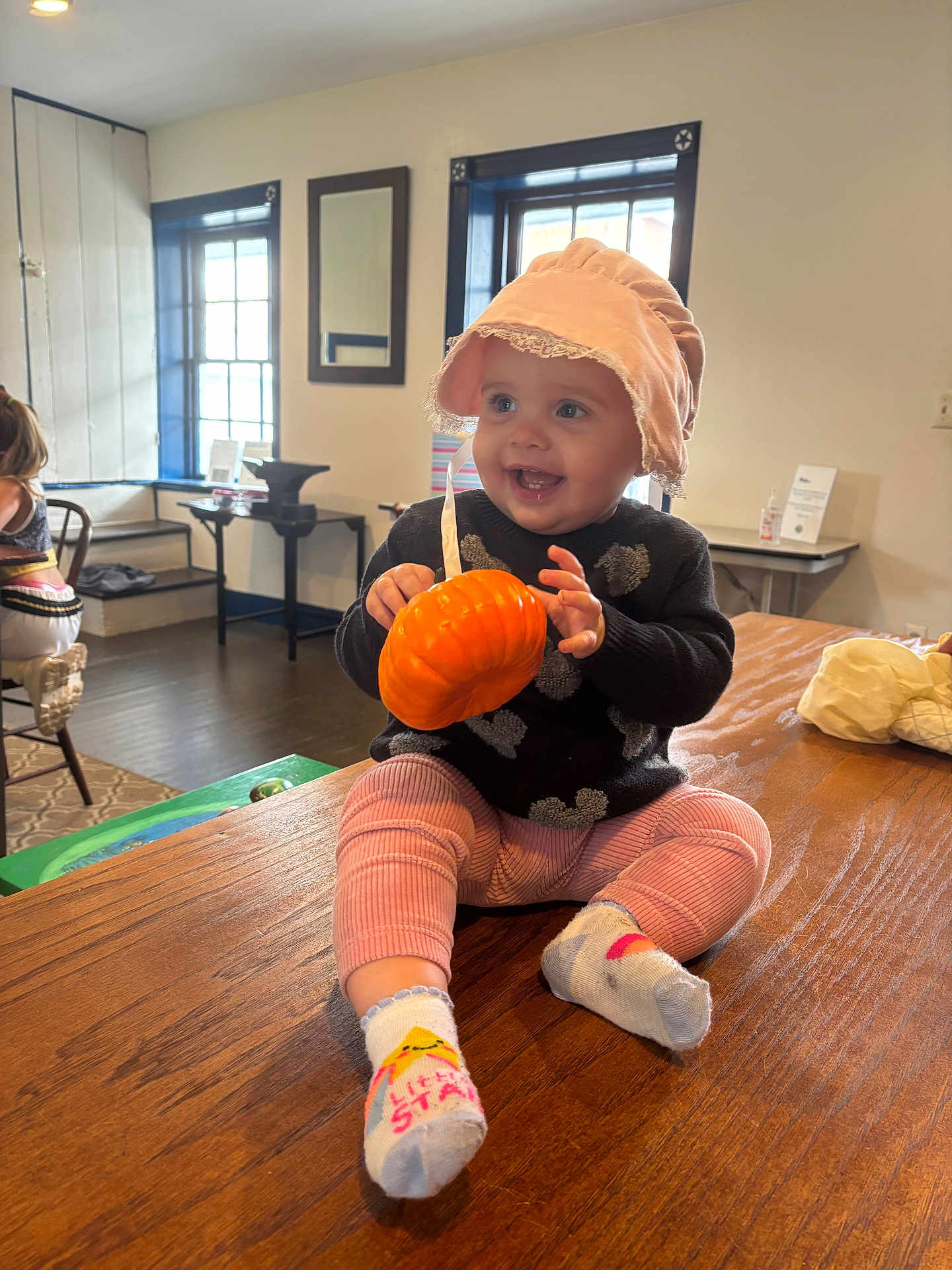 Avis is registered to the contest to win money with this photo: baby, child, smiling, sitting, hat, bonnet, toy, pumpkin, indoor, table, wood, pink_pants, socks, face, person, window, furniture, room, happy, cozy