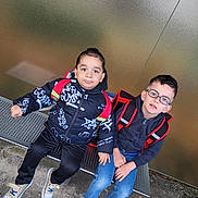 Zyon participe au concours pour gagner de l'argent avec cette photo : child, children, boy, bench, backpack, glasses, jacket, jeans, sneakers, outdoor, casual_clothing, metal_bench, concrete_ground, wall, portrait, two_children, friends, sitting, daylight, urban
