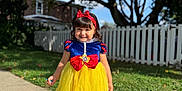 Paulette is registered to the contest to win money with this photo: child, girl, costume, snow_white, dress, yellow_skirt, red_bow, headband, black_shoes, sidewalk, grass, fence, tree, outdoor, sunlight, smile, curly_hair, portrait, happy, daytime