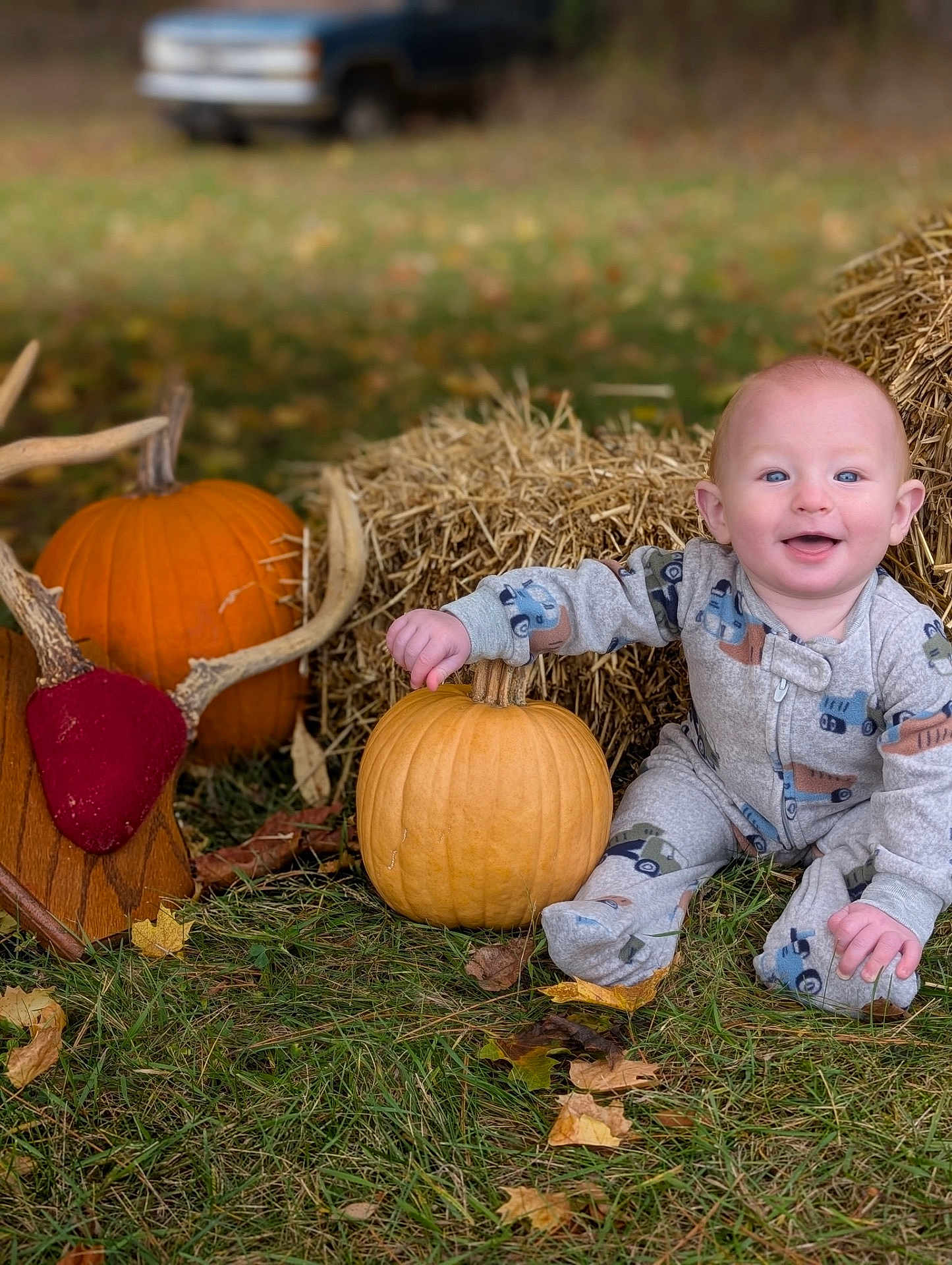 Thad is registered to the contest to win money with this photo: baby, child, pumpkin, hay_bale, grass, leaves, autumn, antler_mount, outdoor, smiling, pajamas, fall, seasonal, decor, nature, person, sitting, blue_eyes, cute, rustic