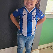 Itzel is registered to the contest to win money with this photo: child, smiling, standing, jeans, sneakers, indoor, window, soccer_jersey, blue, white, hands_on_hips, tile_floor, hair_bow, casual_clothing, portrait, happy, person, young, confident, wall