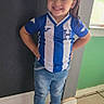 child, smiling, standing, jeans, sneakers, indoor, window, soccer_jersey, blue, white, hands_on_hips, tile_floor, hair_bow, casual_clothing, portrait, happy, person, young, confident, wall