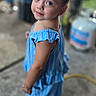 child, girl, blue_eyes, smile, blue_outfit, braided_hair, bracelet, necklace, standing, portrait, outdoor, concrete_floor, blurred_background, casual, cute, person, fashion, young_child, happy, looking_back