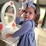 toddler, child, girl, blue_dress, headband, bow, vanity_table, mirror, heart_decorations, indoor, curly_hair, long_sleeves, furniture, playroom, portrait, smiling, cute, young_child, decor, person