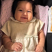 Alaia is registered to the contest to win money with this photo: baby, infant, smile, flower_hair_clips, blanket, pink, dress, knitwear, hand, face, cute, child, soft_lighting, indoors, portrait, happy, lying_down, skin, young_child, cozy