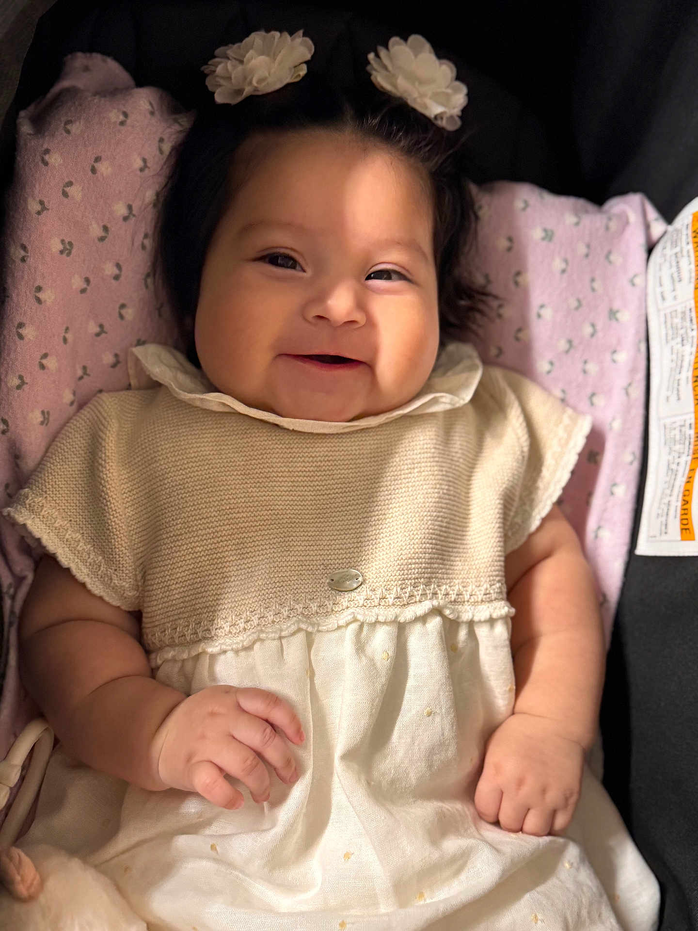 Alaia is registered to the contest to win money with this photo: baby, infant, smile, flower_hair_clips, blanket, pink, dress, knitwear, hand, face, cute, child, soft_lighting, indoors, portrait, happy, lying_down, skin, young_child, cozy