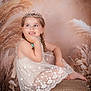 child, girl, smiling, tiara, lace_dress, braided_hair, stool, rustic, pampas_grass, floral_background, portrait, studio_photo, happy, cute, barefoot, decor, seated, young_child, pose, fashion