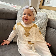 Ayla participe au concours pour gagner de l'argent avec cette photo : child, baby, smiling, headband, couch, pillow, blanket, living_room, white_dress, gold_trim, portrait, happy, sitting, cheeks, hands, eyes, indoors, cozy, candid, infant