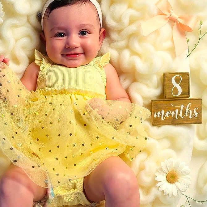Amiliana is registered to the contest to win money with this photo: baby, child, yellow_dress, headband, white_bow, fluffy_blanket, wooden_blocks, flowers, ribbons, smiling, happy, infant, cute, portrait, laying_down, soft_texture, decorative, indoors, skin, legs