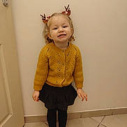 Mya a rejoint le concours — aidez-le/la à gagner de superbes lots ! child, girl, smile, standing, indoor, knitted_sweater, mustard_yellow, black_skirt, black_tights, reindeer_antlers, festive, holiday_accessory, curly_hair, blonde_hair, tile_floor, door, plain_wall, cute, happy, portrait