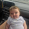 baby, child, infant, person, face, portrait, car_interior, dashboard, clothing, onesie, sun, clouds, arms, hand, seat, seatbelt, window, road, sky, expression