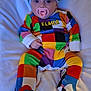 baby, pacifier, colorful_clothing, hat, infant, blanket, lying_down, indoors, cute, patchwork, soft, child, person, cozy, wearing, footed_pajamas, headwear, expression, comfort, young