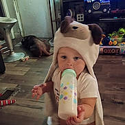 Cheyenne is registered to the contest to win money with this photo: toddler, baby, bottle, hooded_towel, dog, floor, toy, living_room, child, furniture, television, blanket, person, wooden_floor, indoor, cute, animal, home, carpet, sitting