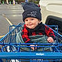 Easton is registered to the contest to win money with this photo: baby, shopping_cart, beanie, red_and_black_clothing, blanket, parking_lot, smile, face, child, outdoor, toy, vehicle, person, winter_clothing, cute, expression, seat, hand, footwear, daylight