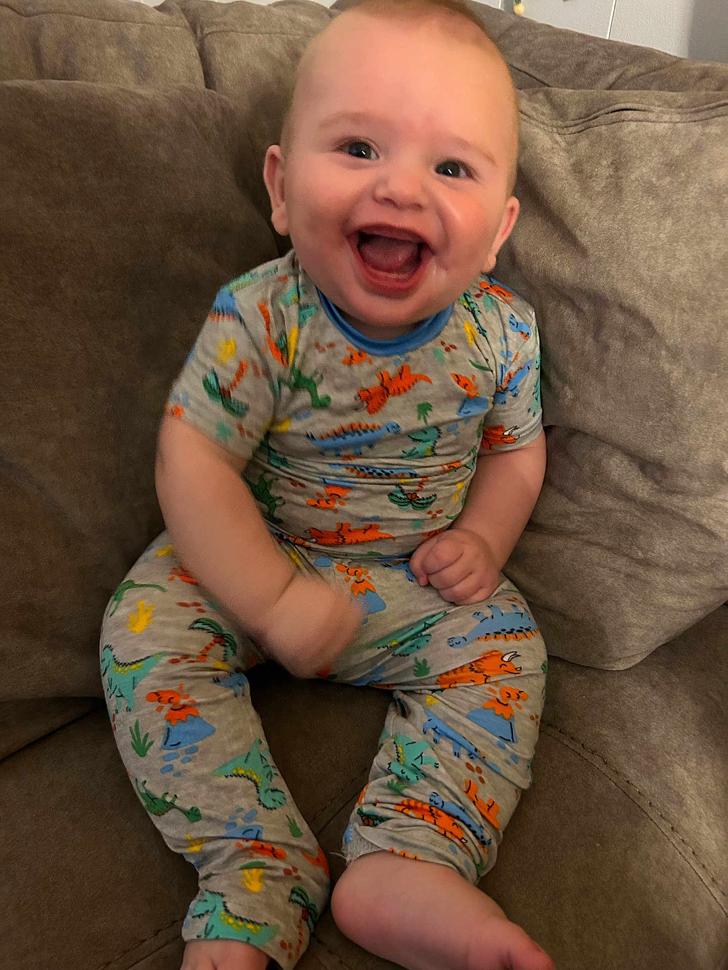 Easton joined the competition — help win amazing prizes! baby, child, smiling, laughing, pajamas, dinosaur_print, couch, indoor, happy, person, sitting, cute, infant, cozy, comfortable, clothing, living_room, young, portrait, playful