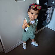 Kevin participe au concours pour gagner de l'argent avec cette photo : toddler, child, kitchen, overalls, sneakers, sunglasses, snack, standing, floor, dishwasher, oven, white_shirt, chain_necklace, curly_hair, expression, indoor, appliance, clothing, person, footwear