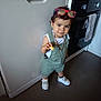 toddler, child, kitchen, overalls, sneakers, sunglasses, snack, standing, floor, dishwasher, oven, white_shirt, chain_necklace, curly_hair, expression, indoor, appliance, clothing, person, footwear
