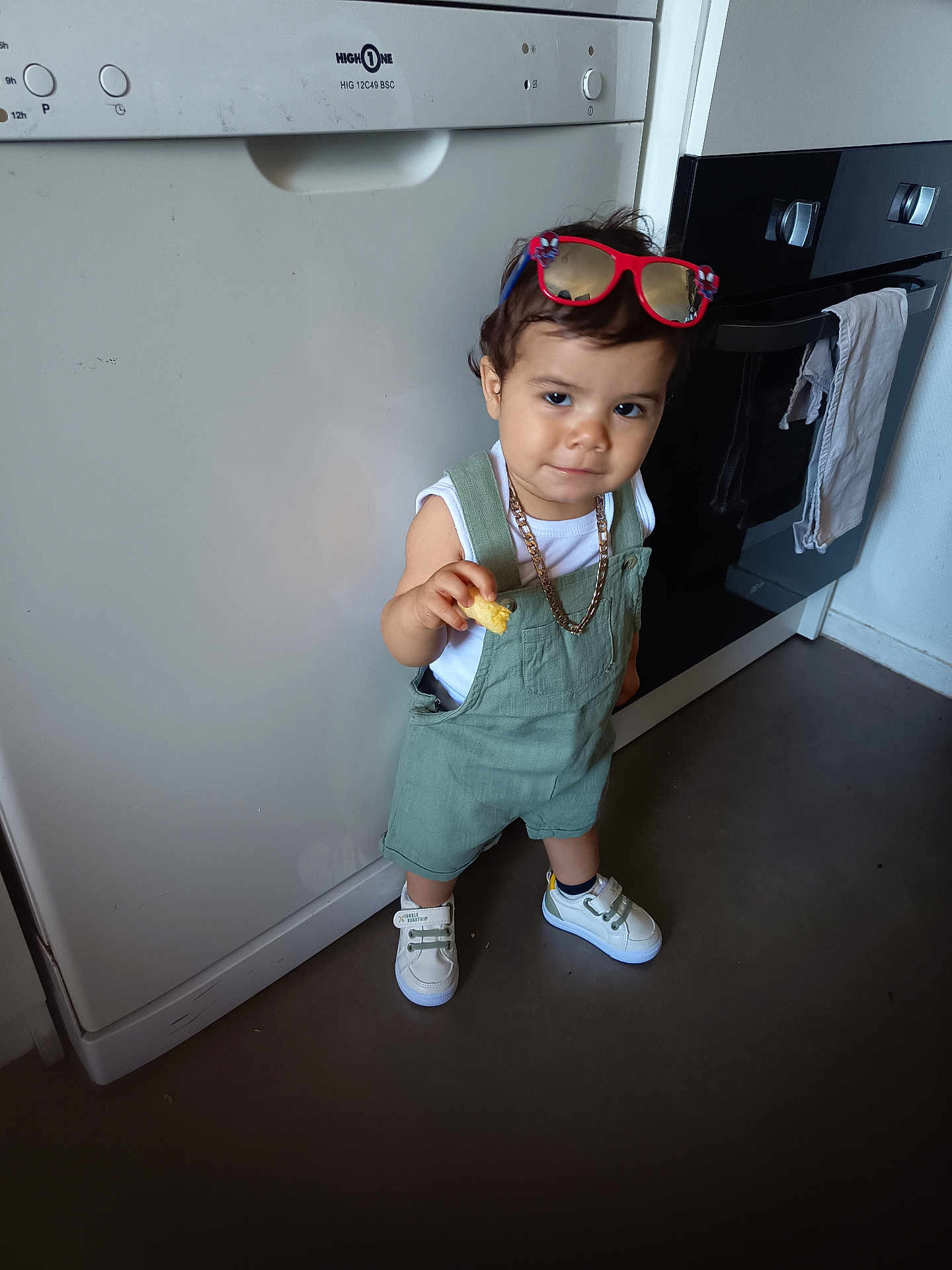 Kevin participe au concours pour gagner de l'argent avec cette photo : toddler, child, kitchen, overalls, sneakers, sunglasses, snack, standing, floor, dishwasher, oven, white_shirt, chain_necklace, curly_hair, expression, indoor, appliance, clothing, person, footwear