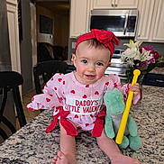 Journee is registered to the contest to win money with this photo: baby, infant, child, headband, red_bow, valentine_outfit, heart_pattern, smile, sitting, countertop, granite_counter, kitchen, microwave, cabinets, plush_toy, green_toy, yellow_stick, flowers, bouquet, bare_feet