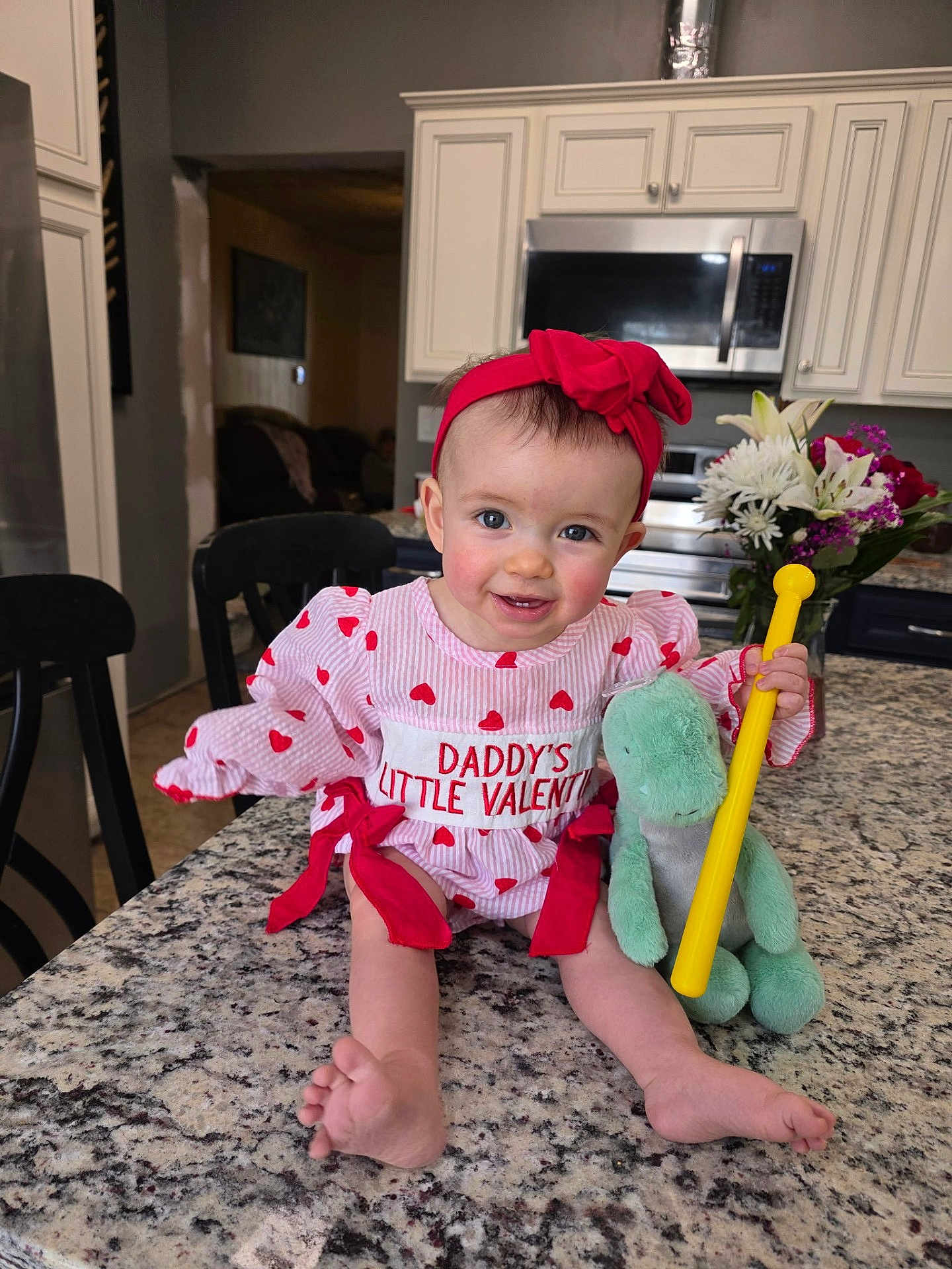 Journee is registered to the contest to win money with this photo: baby, infant, child, headband, red_bow, valentine_outfit, heart_pattern, smile, sitting, countertop, granite_counter, kitchen, microwave, cabinets, plush_toy, green_toy, yellow_stick, flowers, bouquet, bare_feet