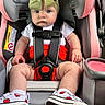baby, child, car_seat, headband, bow, red_shorts, white_shoes, apple_design, seatbelt, straps, serious_expression, infant, cute, sitting, safety, pink, gray, clothing, footwear, indoors
