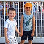 child, boy, smiling, holding_hands, outdoor, fence, cap, tshirt, shorts, dinosaur_print, sunny, people, background, happy, casual_clothing, summer, friends, playful, portrait, street