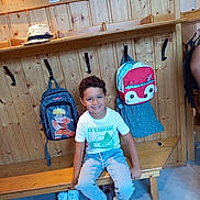 Taim a rejoint le concours — aidez-le/la à gagner de superbes lots ! child, boy, smile, bench, backpack, wooden_wall, wooden_bench, shoes, casual_clothing, curly_hair, indoor, tile_floor, hooks, seat, happy, white_tshirt, blue_jeans, playful, school_bag, portrait
