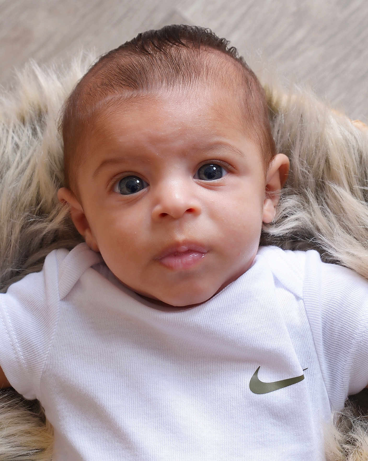 Kameron joined the competition — help win amazing prizes! baby, infant, face, eyes, clothing, white_shirt, nike_logo, fur_blanket, portrait, closeup, cute, newborn, child, young, expression, skin, head, person, laying, soft