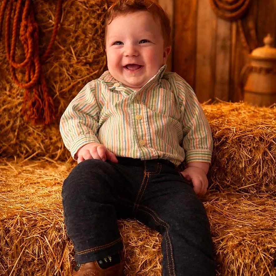 Colbin Shean joined the competition — help win amazing prizes! toddler, child, smiling, hay_bales, cowboy_hat, rope, barn, rustic, jeans, striped_shirt, boots, wooden_wall, string_lights, indoors, portrait, happy, seated, warm_lighting, farm, clothing