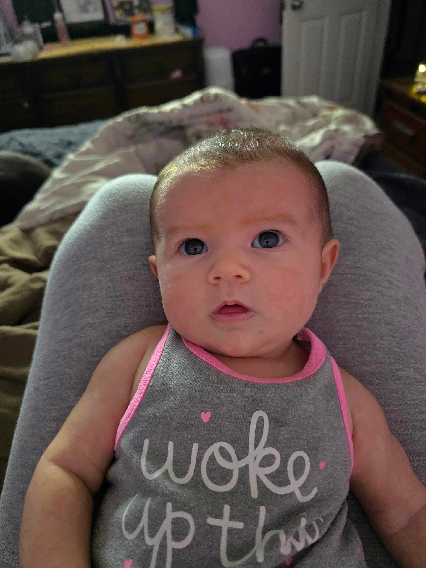 Isabella joined the competition — help win amazing prizes! baby, infant, face, person, clothing, tank_top, pink, gray, eyes, arms, indoor, bedroom, blanket, furniture, curious, sitting, skin, head, expression, human