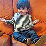 toddler, child, striped_shirt, jeans, sitting, orange_couch, indoor, casual_clothing, curious_expression, small_object, footwear, young_child, portrait, home_interior, comfortable, cute, person, furniture, relaxed, seated