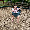 baby, swing, playground, child, smiling, happy, outdoor, woodchips, greenery, tree, fence, clothing, person, seat, fun, nature, toy, baby_clothes, summer, park