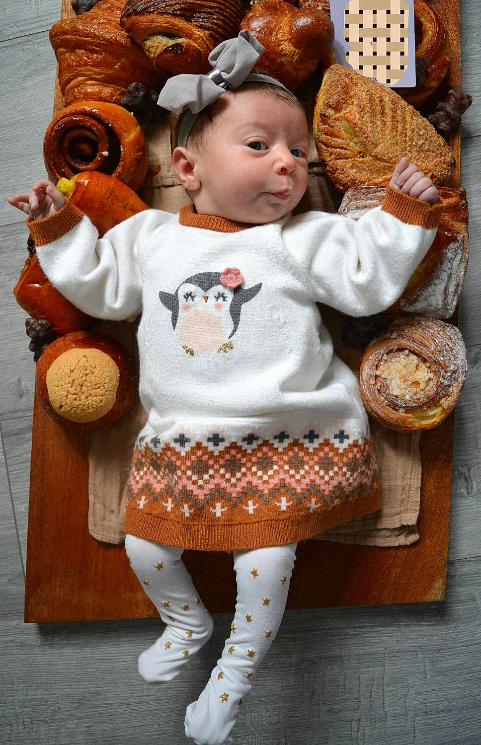 Hana participe au concours pour gagner de l'argent avec cette photo : baby, infant, child, pastries, bread, croissant, cinnamon_roll, wooden_board, sweater, headband, penguin_design, leggings, floor, food, breakfast, cute, cozy, expression, lying_down, indoors