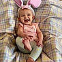 baby, child, bunny_ears, pink_clothing, smiling, playful, blanket, colorful, patterned, infant, happy, cute, indoors, person, legs, feet, headband, sitting, joyful, portrait