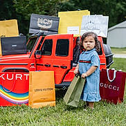 Lia is registered to the contest to win money with this photo: child, curly_hair, cute, daylight, denim_dress, fashion, grass, green_grass, luxury_brands, outdoor, person, play, red_car, retail, serious_expression, shopping_bags, standing, summer, toddler, toy_car