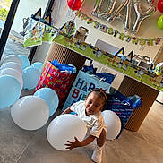 Khyree is registered to the contest to win money with this photo: toddler, balloon, birthday_party, gift_bag, indoor, child, celebration, decorations, white_balloon, colorful_balloons, party_table, floor, casual_clothing, happy, smiling, braided_hair, shoes, present, wall_decor, festive